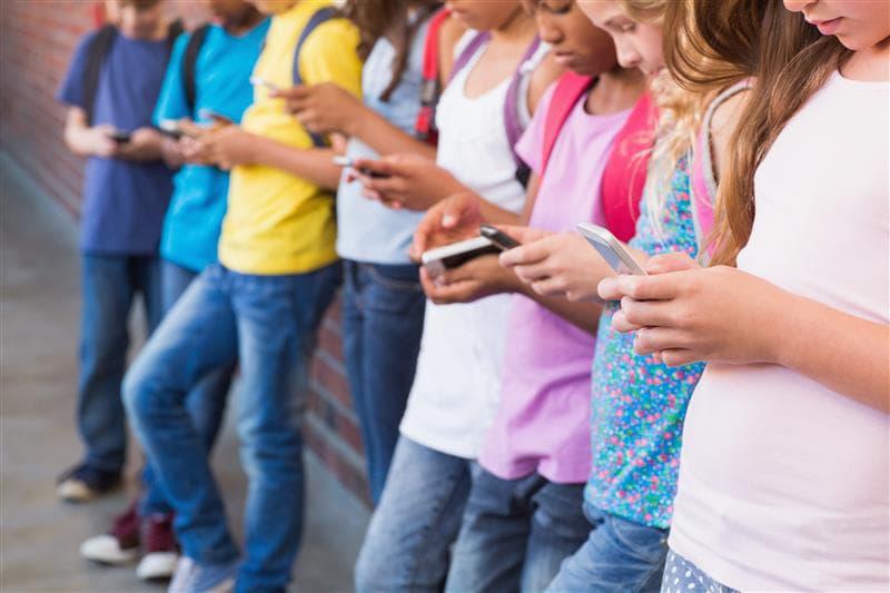 young students leaning against a wall using cell phone devices